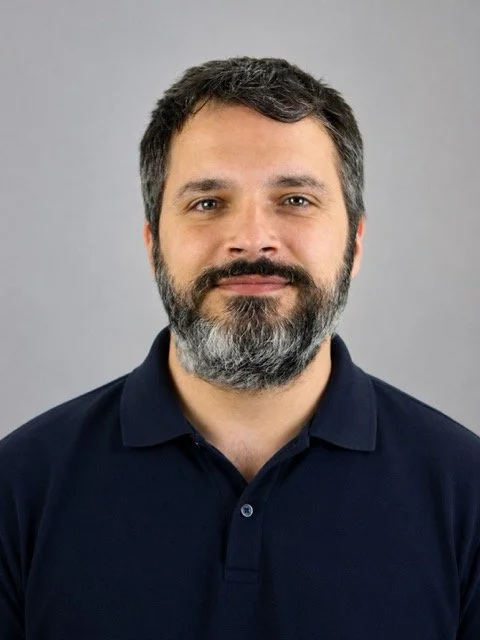 Portrait of a man with dark hair, a full beard, wearing a navy blue polo shirt, smiling slightly, against a plain gray background.
