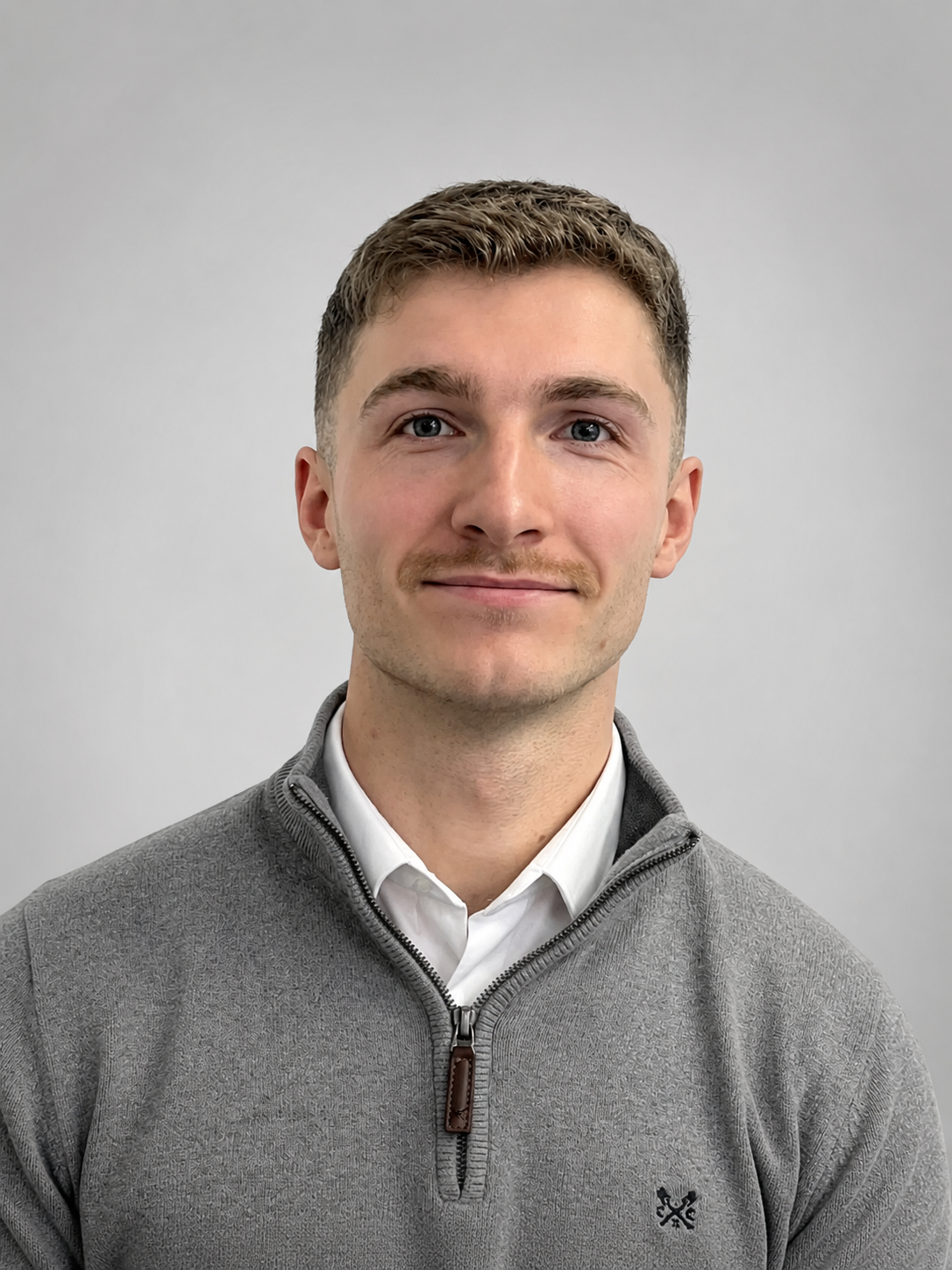 Portrait of a young man with short brown hair, blue eyes, and light beard, smiling, wearing a gray zip-up sweater over a white collared shirt, against a plain light gray background.