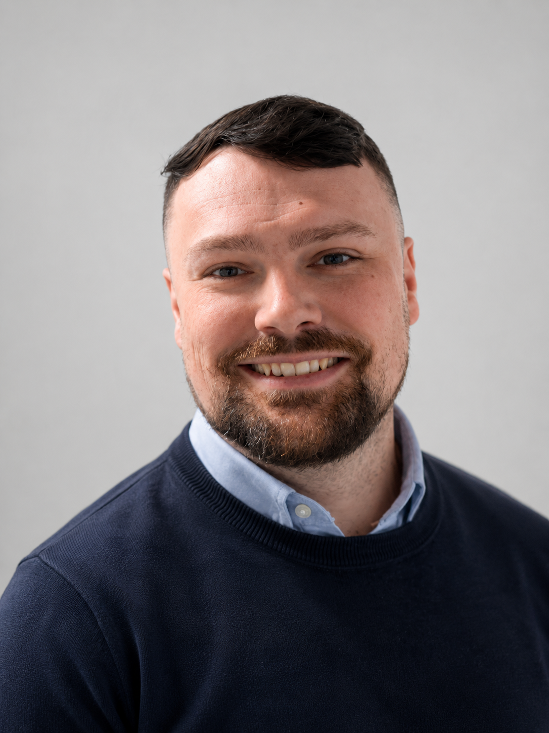 A portrait of a smiling man with short dark hair, a beard, and blue eyes, wearing a navy sweater over a light blue collared shirt, against a plain light gray background.