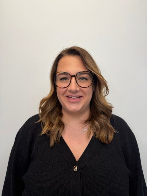 A woman with shoulder-length wavy brown hair, glasses, and a black cardigan standing against a plain white wall.
