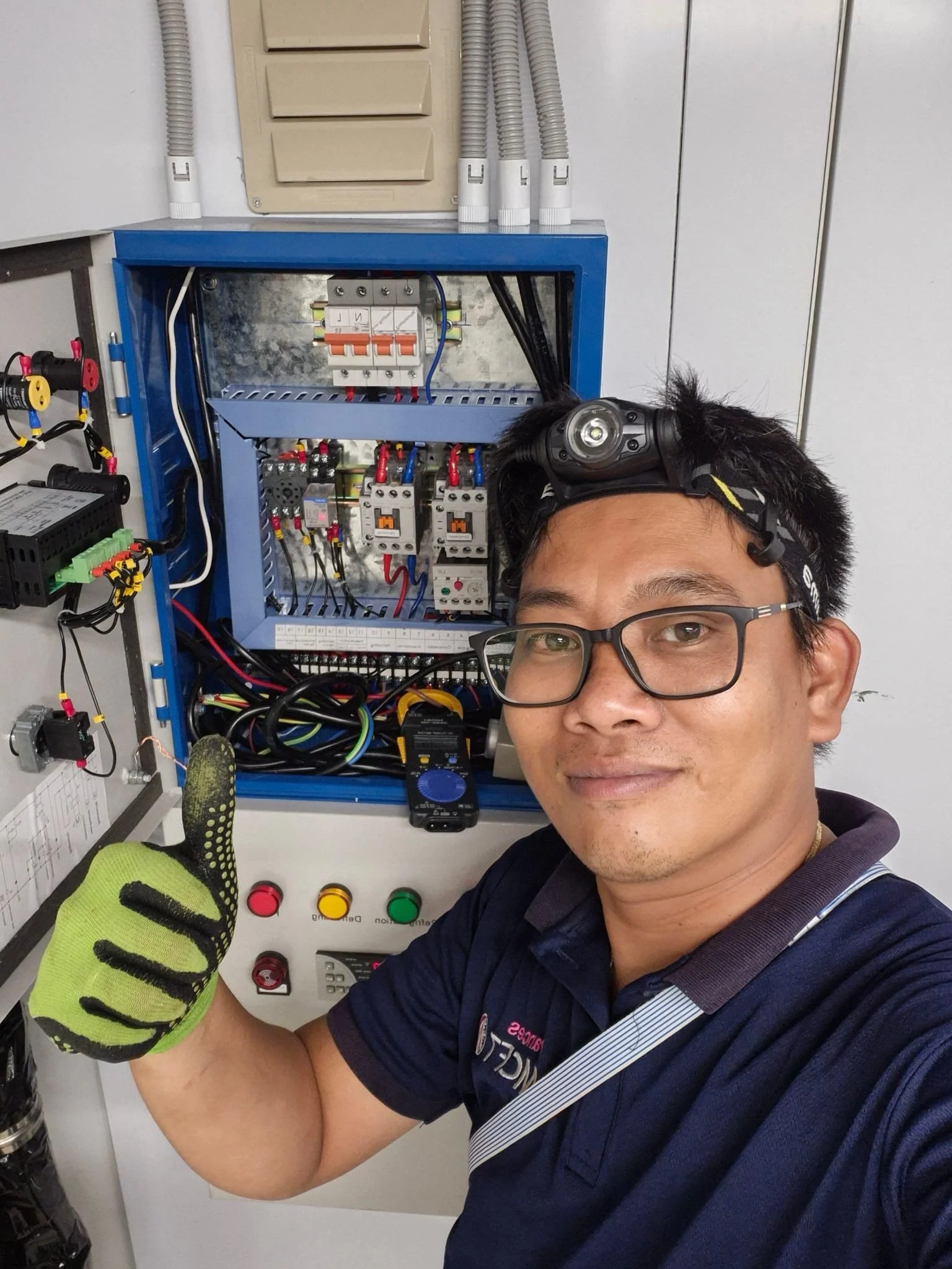 A smiling man wearing glasses and a headlamp, giving a thumbs up with his left hand, wearing a navy blue work shirt, standing in front of an electrical panel with wiring and circuit breakers, in an indoor setting.