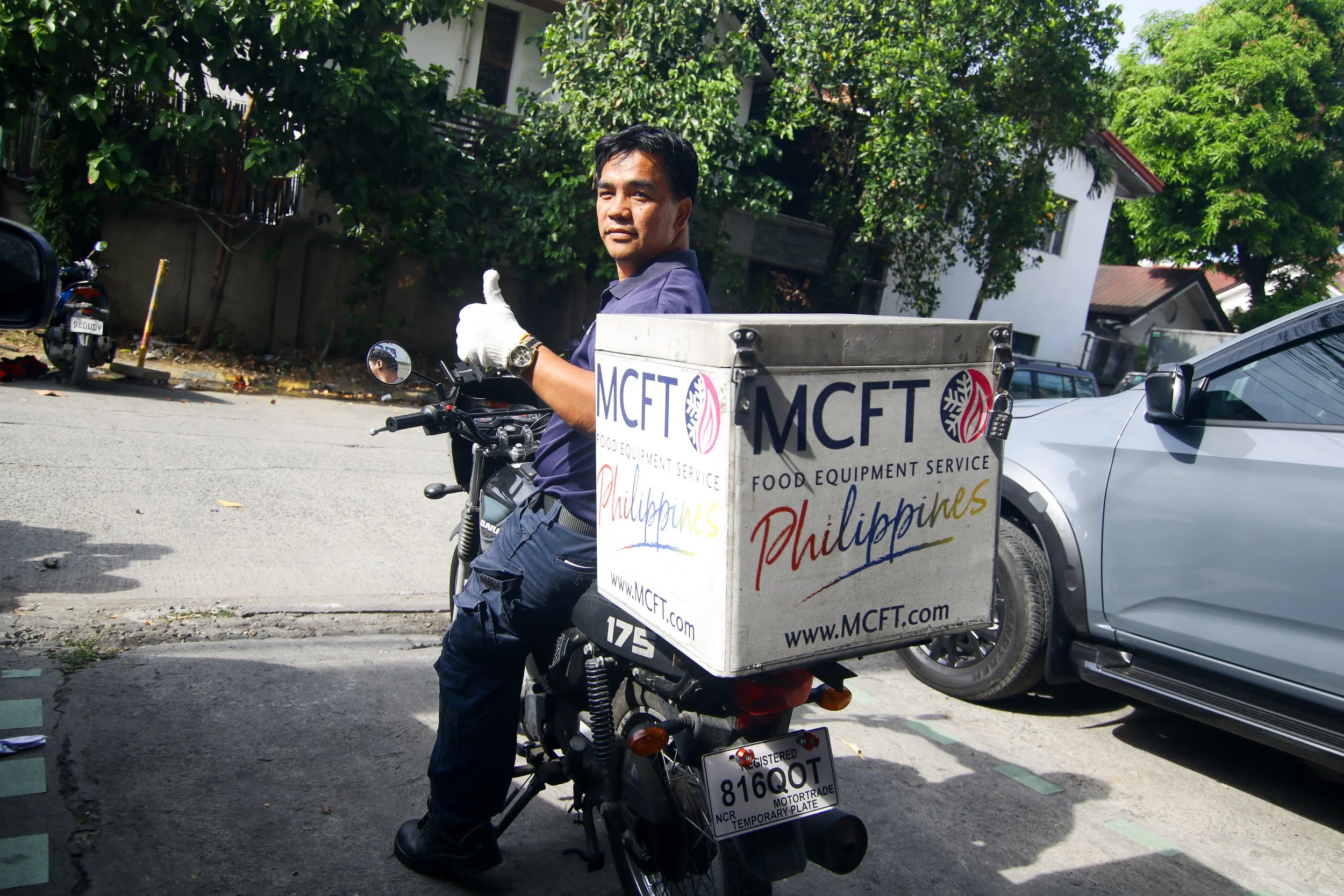 Man riding a motorcycle, giving a thumbs-up, with a box attached to the back that has 'MCFT Food Equipment Service Philippines' written on it. He is on a street with parked cars, greenery, and houses in the background.