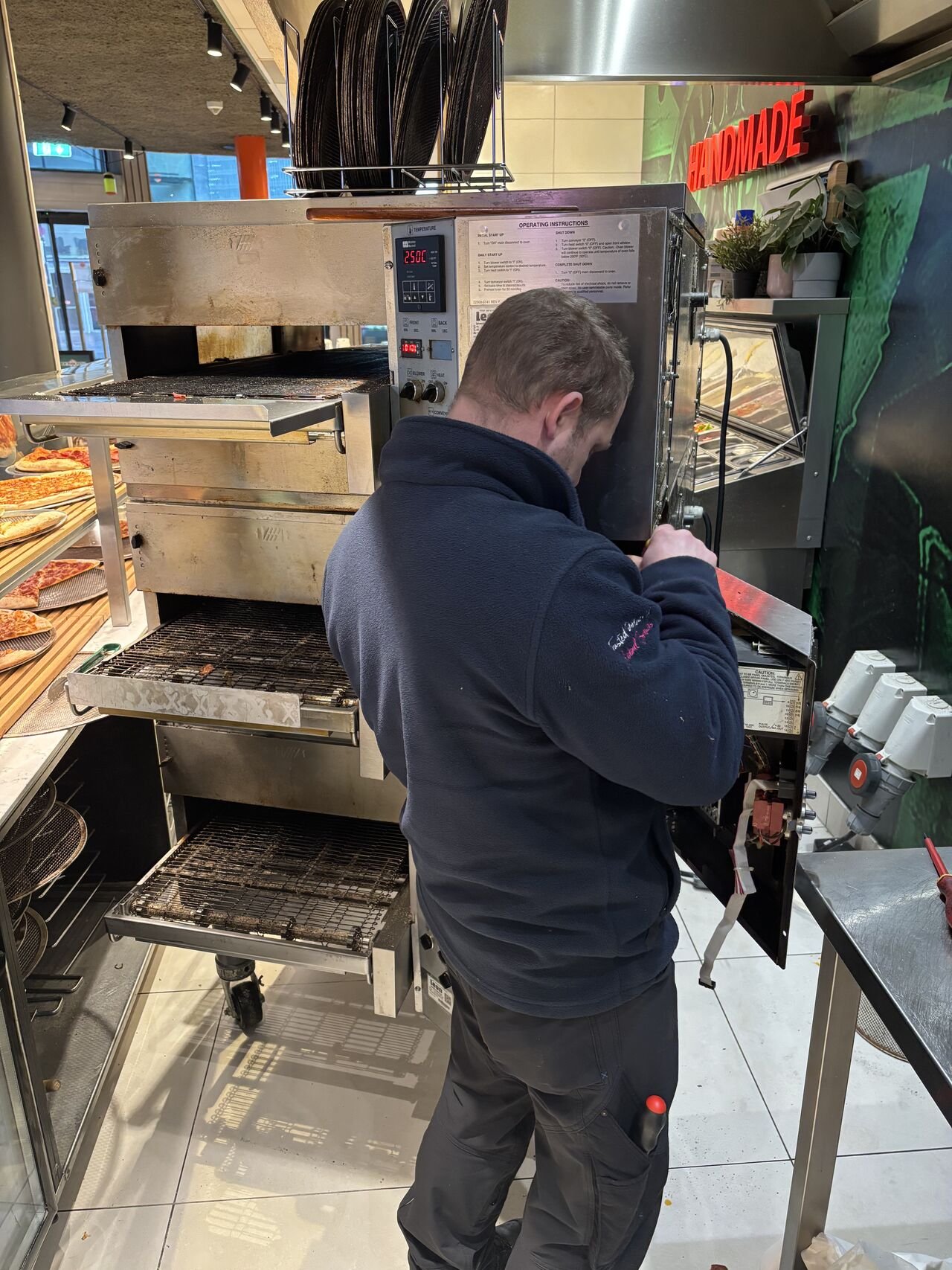 A man with short hair wearing a black jacket is looking down and working on a large piece of equipment, which appears to be an oven or bakery machine, in a commercial kitchen or bakery.