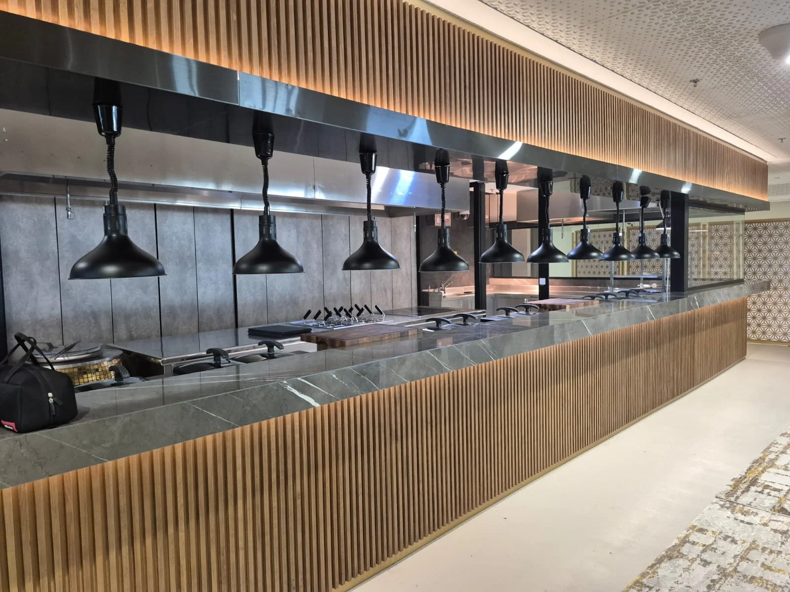A modern buffet counter with a marble surface and wooden vertical slats underneath, featuring black hanging lamps above and a gray tiled backsplash, in a well-lit indoor setting.