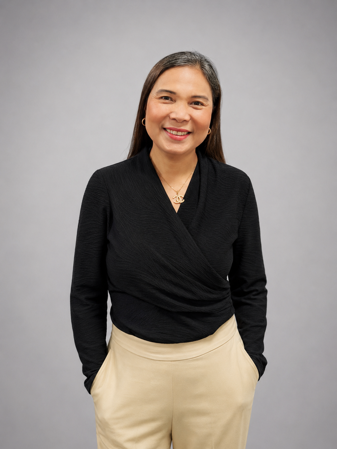 A woman with long dark hair smiling, wearing a black textured wrap top, beige pants, gold jewelry, and standing against a plain light gray background.
