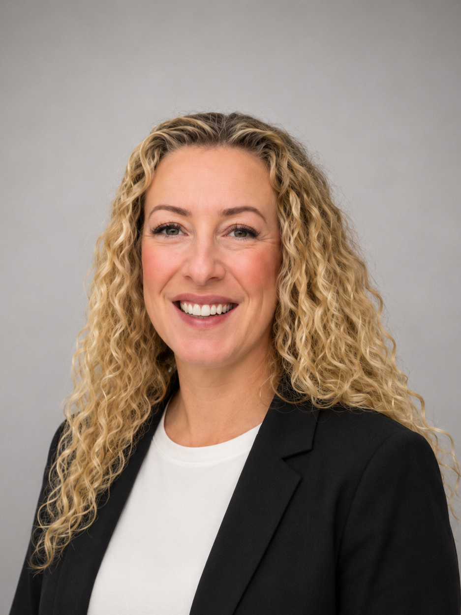 A woman with long, curly blonde hair smiling at the camera, wearing a black blazer over a white shirt, against a gray background.
