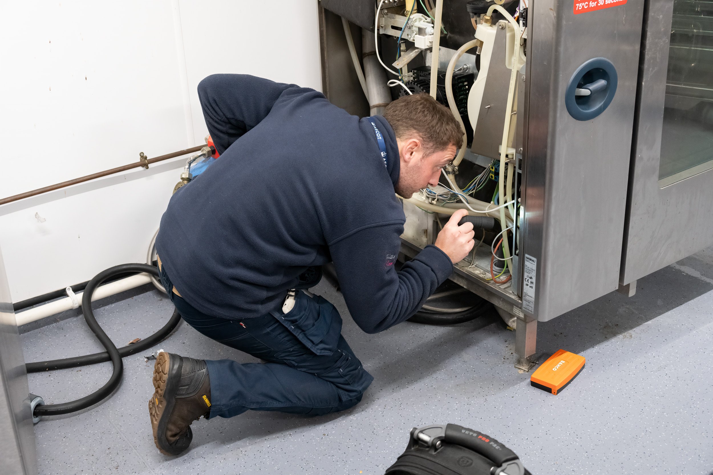 A man crouches down working on an open industrial machine with electrical and plumbing components. He is wearing a dark jacket, jeans, and work boots, and is holding a flashlight to examine the internal parts. The setting appears to be a workshop or maintenance area with gray floors and a white wall.