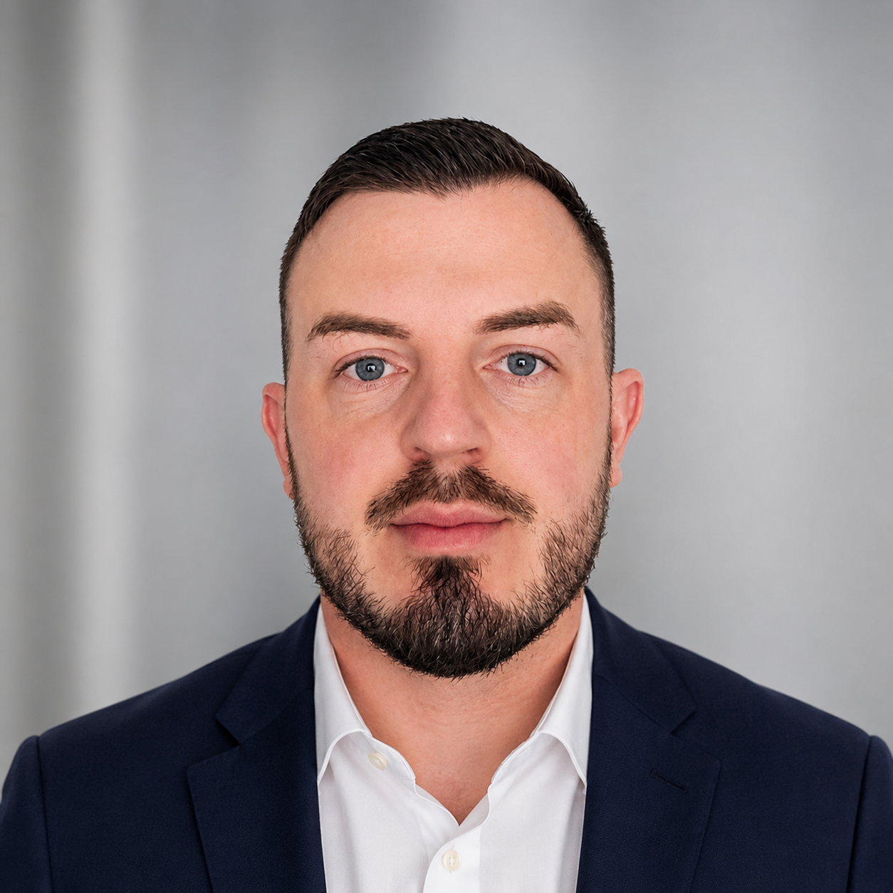 Portrait of a man with blue eyes, short dark hair, and a beard, wearing a white shirt and navy blazer against a gray background.