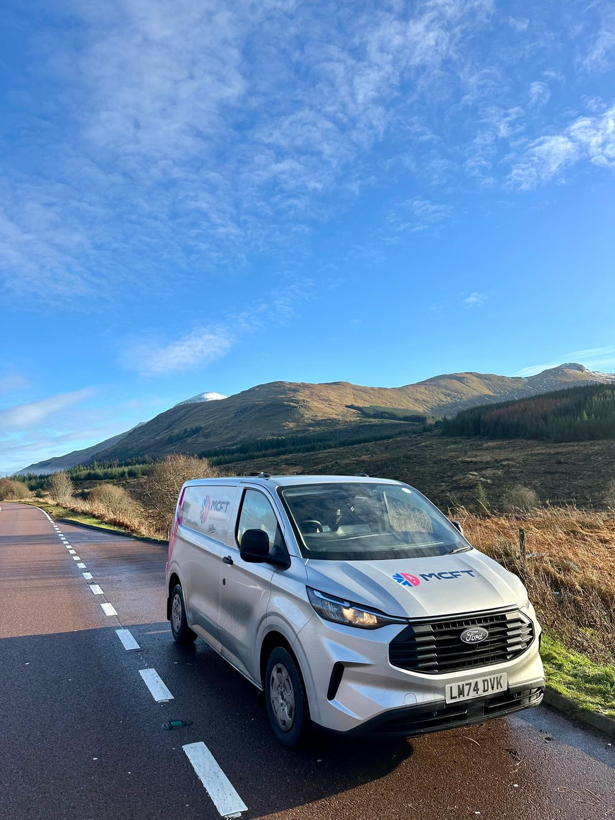 A white Ford Transit Connect van with 'MCFT' logo parked on the side of a scenic mountain road with mountains and blue sky in the background.