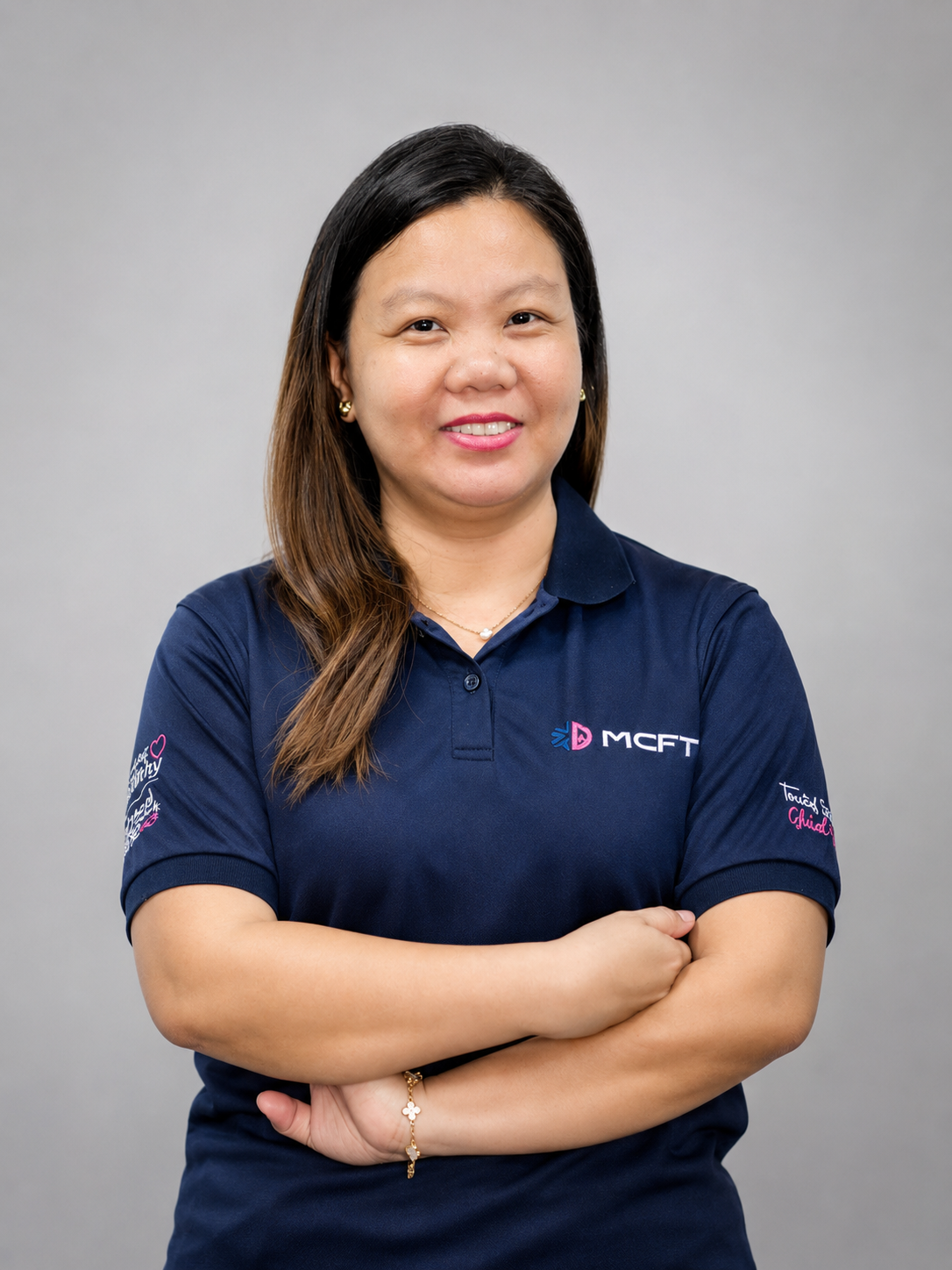 A woman with long brown hair, wearing a navy blue polo shirt with logos, standing with arms crossed against a plain gray background.