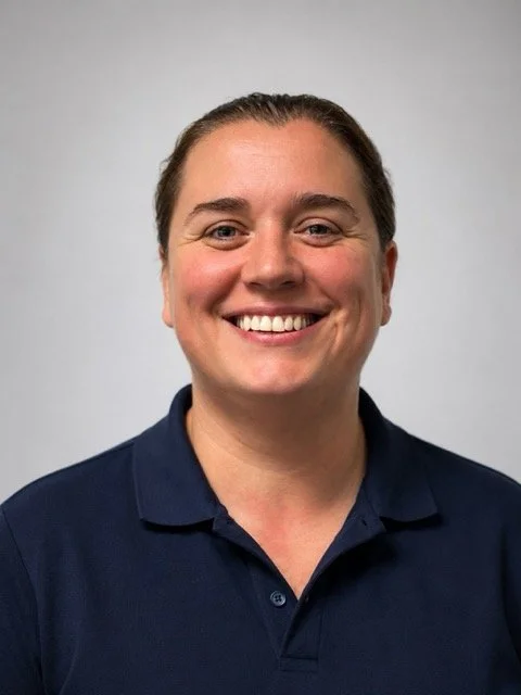 A woman with brown hair pulled back, smiling, wearing a navy blue polo shirt, against a plain gray background.