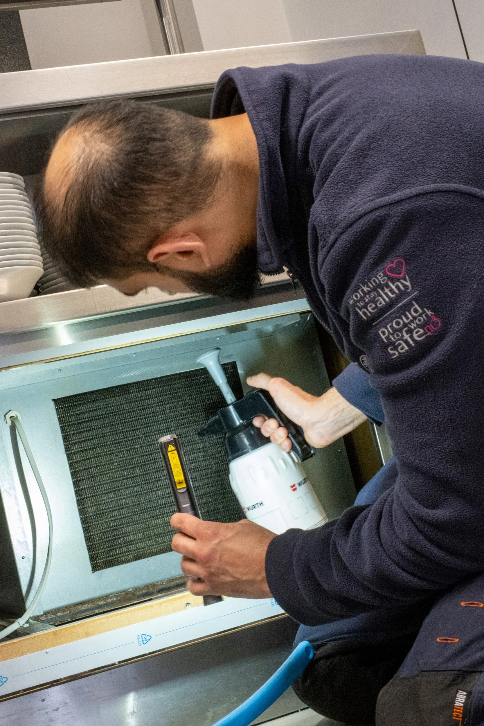 A person wearing a navy blue fleece jacket with a pink heart and text, working on installing or repairing an HVAC unit or air conditioning system in a kitchen area.