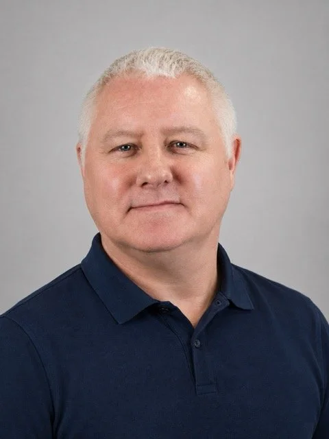 A middle-aged man with short, white hair and light skin, wearing a dark blue polo shirt, standing against a plain gray background.