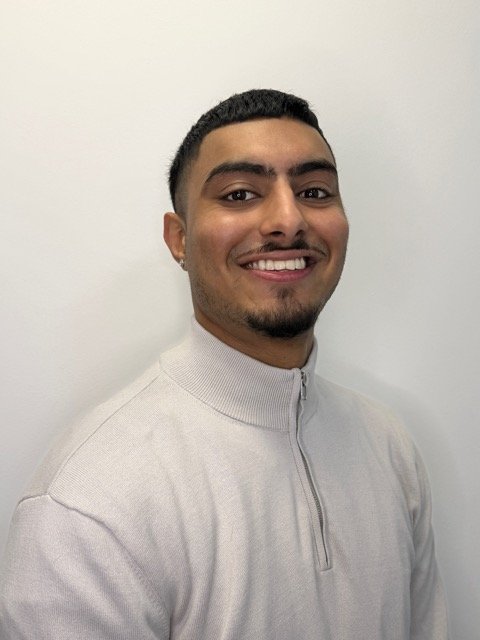 Smiling young man with short dark hair, facial hair, earrings, wearing a light gray zip-up sweater standing against a plain white wall.