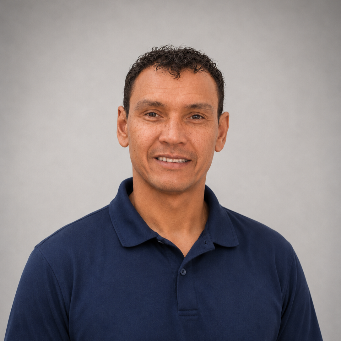 A man with short, curly brown hair wearing a navy blue polo shirt, smiling slightly, standing against a plain light gray background.