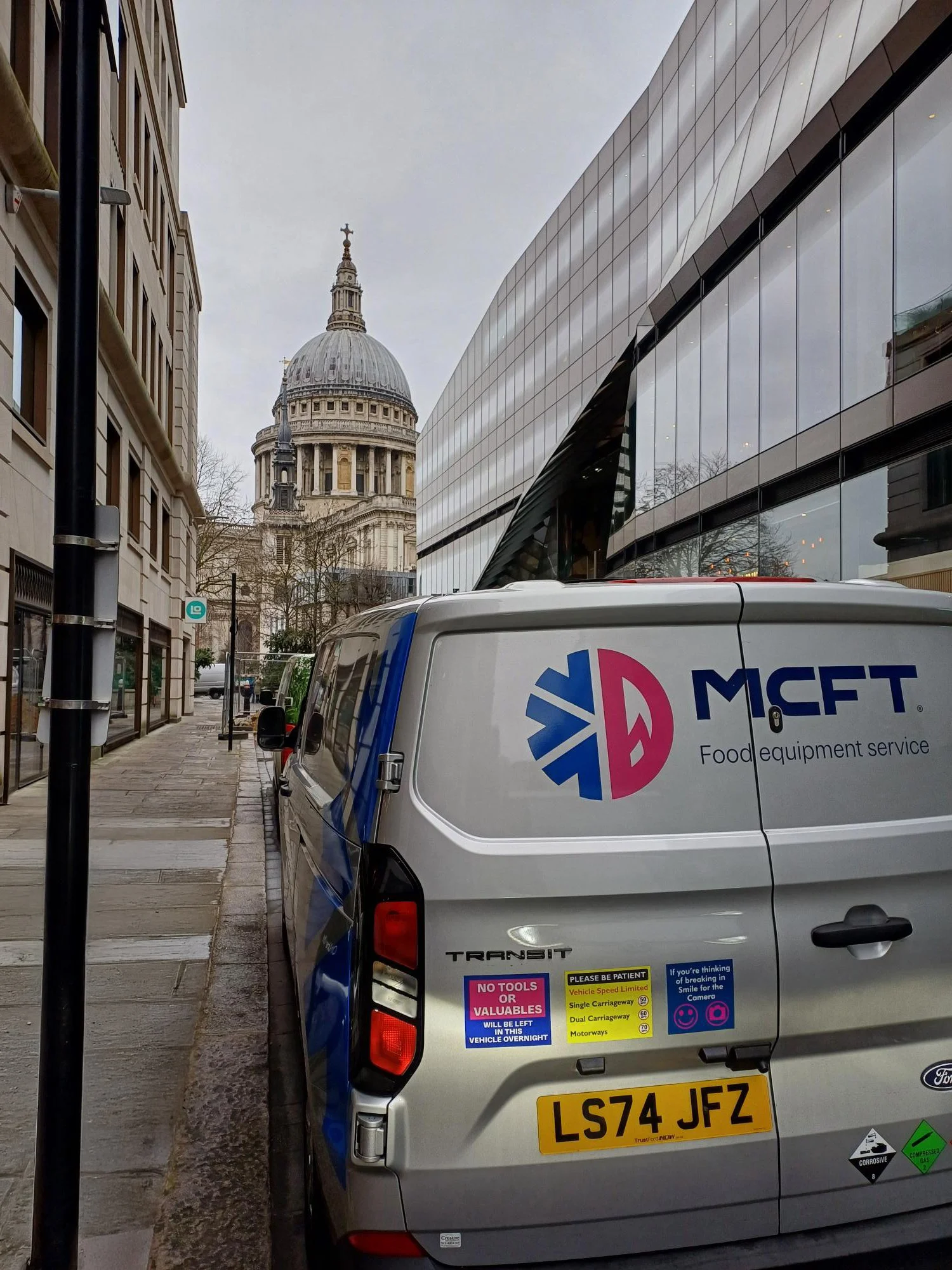 View of a city street with a parking van in the foreground, featuring company branding and safety stickers, and historic landmarks with a dome and cross in the background under an overcast sky.