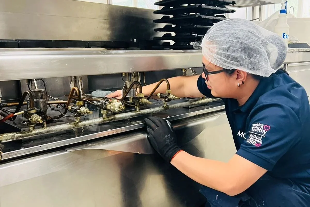 A woman wearing glasses, a hairnet, and a uniform while inspecting or repairing a commercial oven or stove in a kitchen.
