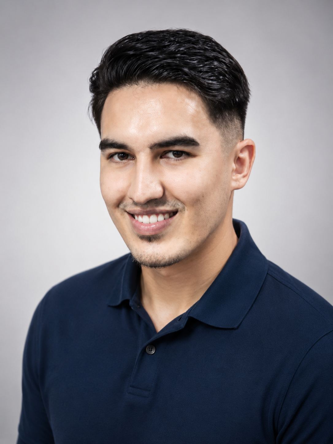 A young man with dark, slicked-back hair, a light complexion, and a trimmed goatee, smiling in a navy blue polo shirt, against a plain gray background.