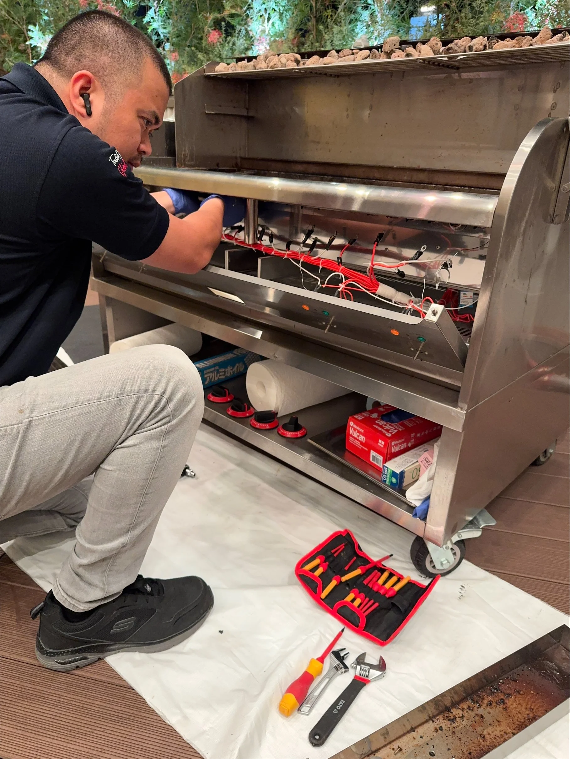 A person kneeling on a protective sheet repairing or maintaining a stainless steel barbecue grill. The person is using tools from a red toolbox which is open on the floor. There are various tools including screwdrivers, a wrench, and pliers visible. The person has black shoes, light-colored pants, and is wearing one black earpiece.