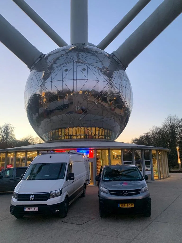 A futuristic building with a large, reflective, spherical structure on top of a circular base, with two cars parked in front, one white van and one black compact car, during dusk.
