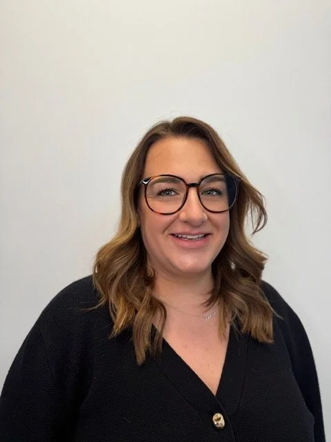 A woman with shoulder-length brown hair, wearing glasses with dark frames and a black top, smiling in front of a plain white wall.