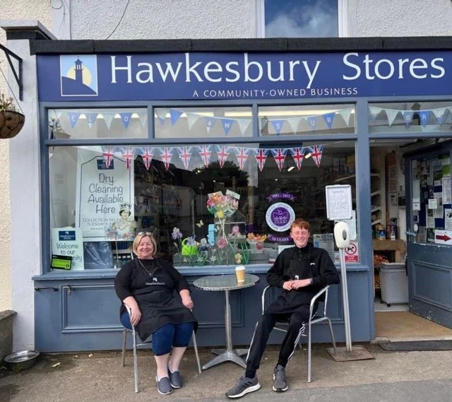 Two people sitting outside a community-owned store called Hawkesbury Stores, with decorations and Union Jack flags in the display window.