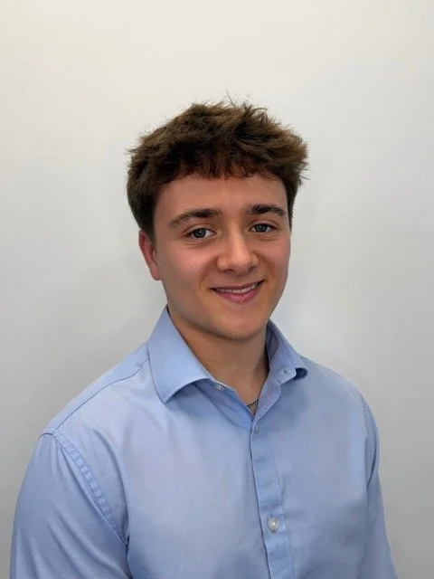 A young man with brown hair, blue eyes, and fair skin, wearing a light blue button-up shirt, smiling at the camera against a plain white wall.