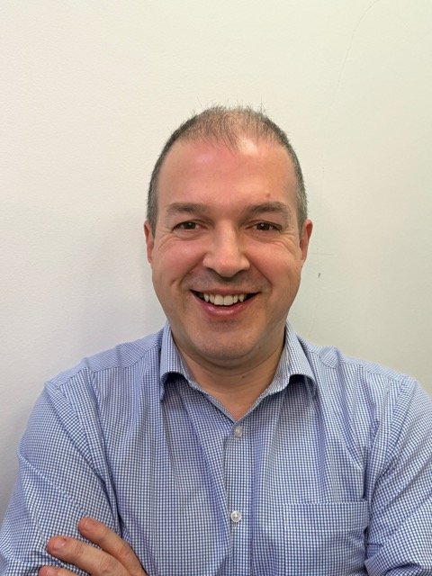 A man with short brown hair, smiling, wearing a blue checkered shirt, standing against a plain light-colored wall.