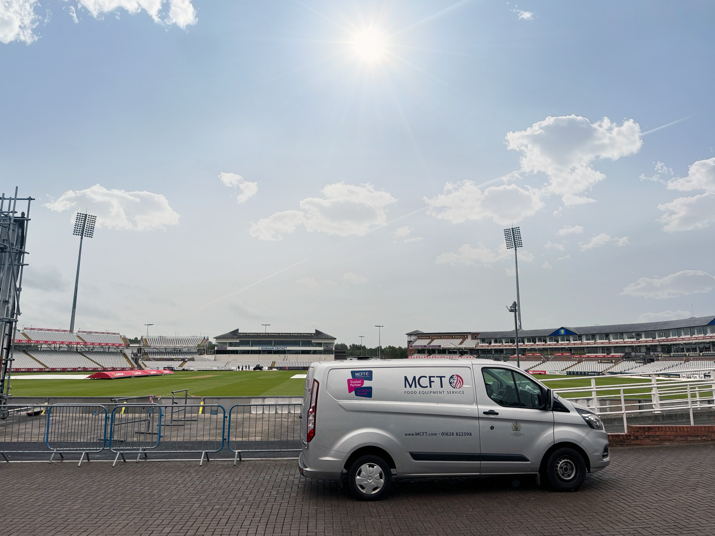 A cricket stadium with empty stands and floodlights, with a partially cloudy sky and the sun shining brightly. A white vans with advertising and a fence are in the foreground.