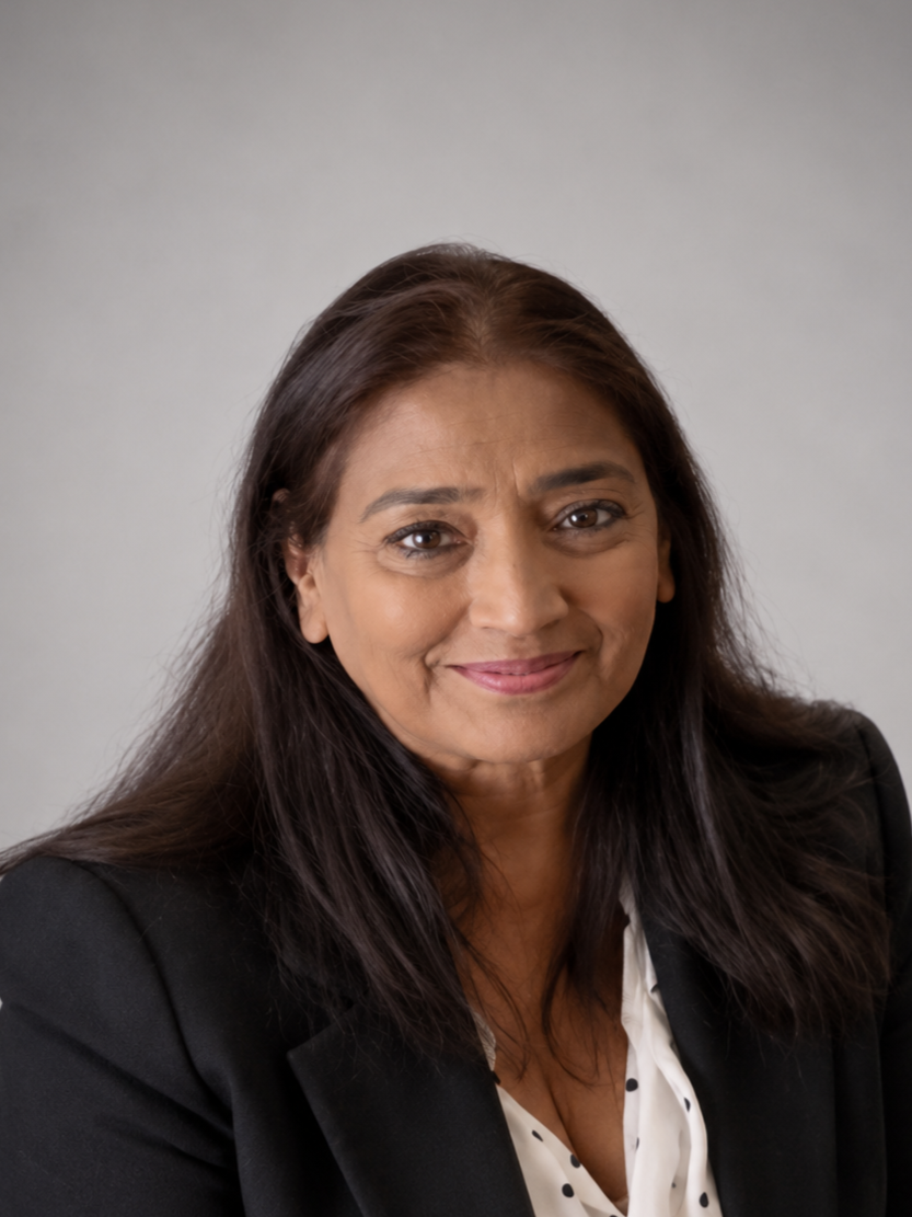 A woman with long dark brown hair wearing a black blazer and white blouse, smiling softly, against a plain light grey background.