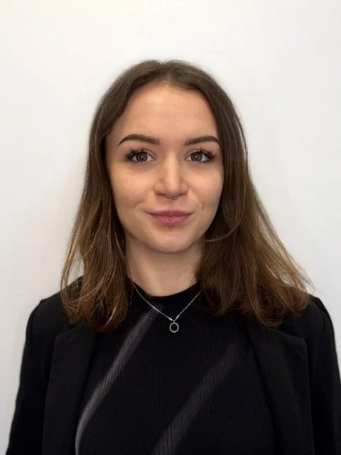 A young woman with shoulder-length brown hair, light skin, and facial piercings posing against a plain white wall, wearing a black top and a delicate necklace.