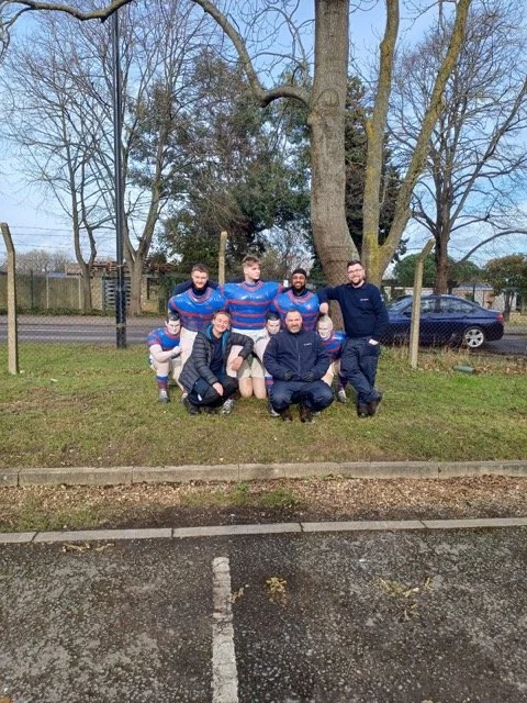 Group of seven people posing outdoors in front of large trees, with some wearing sports uniforms and others in casual clothing.