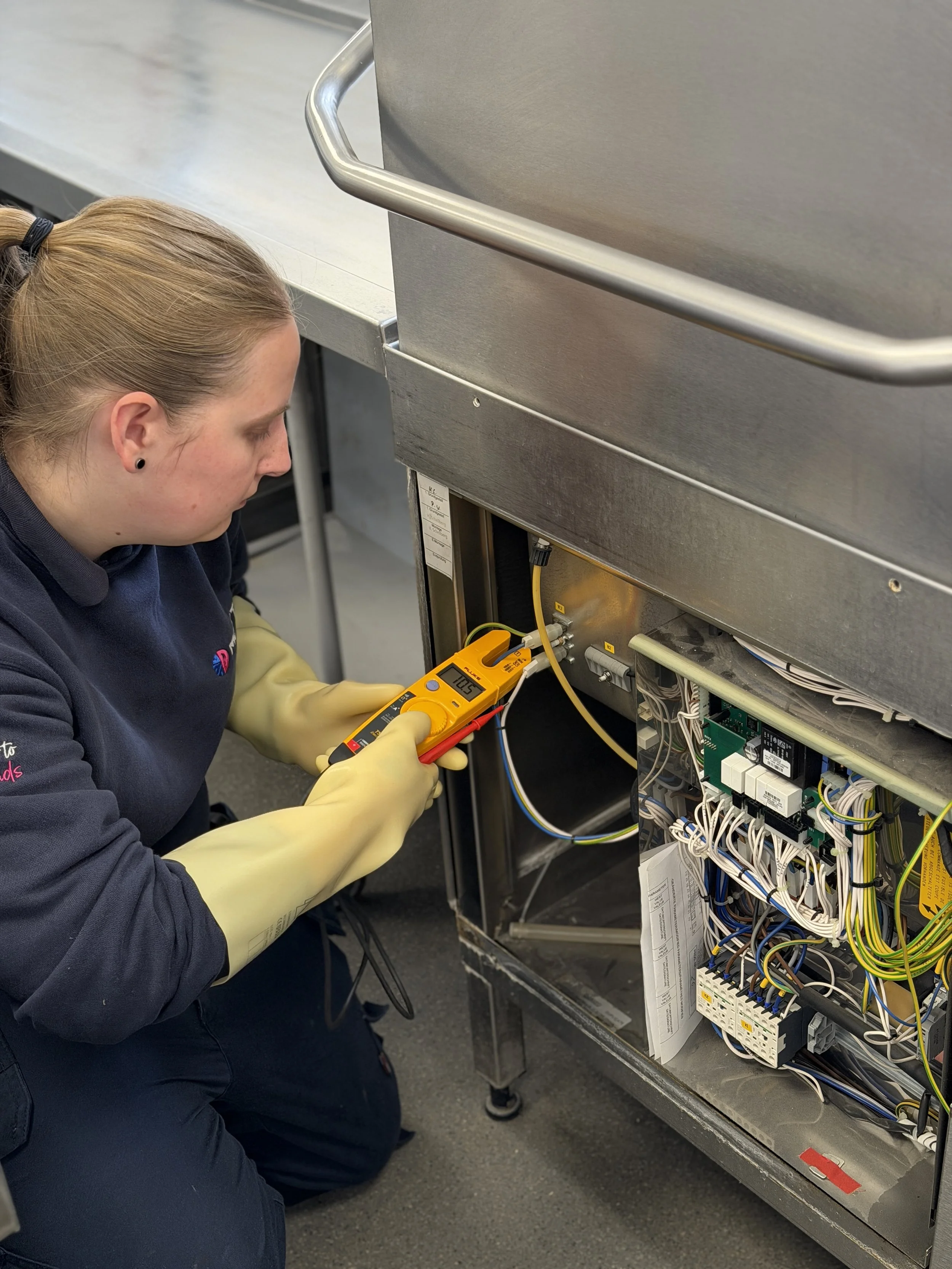 Technician wearing gloves working on electrical or control panel with multimeter in a commercial or industrial setting.