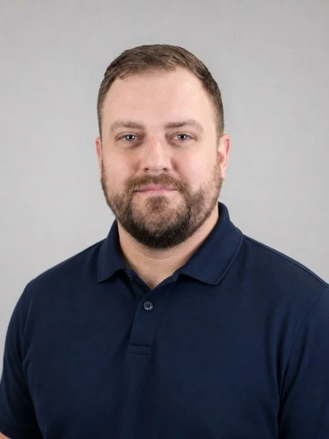 Portrait of a man with short brown hair and beard, wearing a navy blue polo shirt, against a plain gray background.