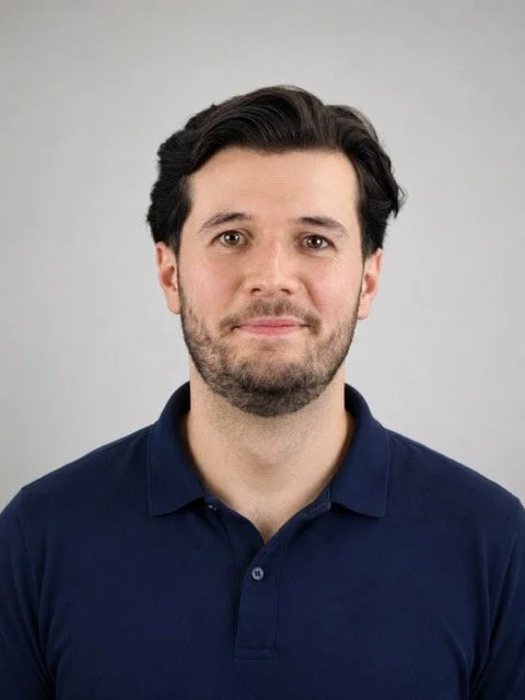 Portrait of a man with dark hair and a beard, wearing a navy blue polo shirt, looking at the camera against a gray background.