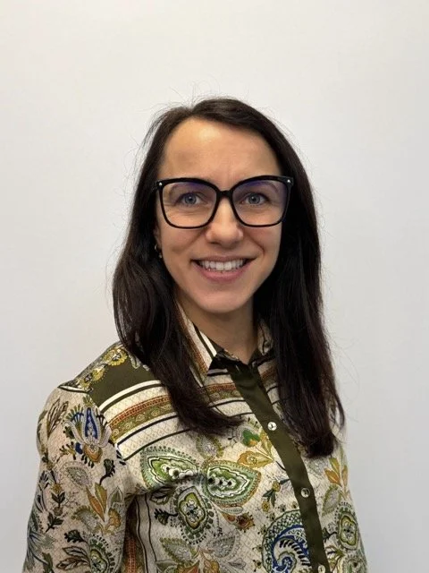 A woman with shoulder-length brown hair, wearing black-rimmed glasses and a patterned blouse, smiling in front of a plain white wall.