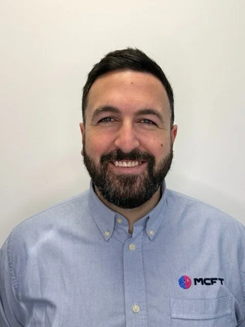 Portrait of a man with dark hair and beard, smiling, wearing a light blue button-up shirt with an MCFT logo on the chest, against a plain off-white background.