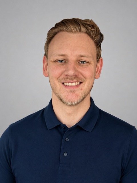 Portrait of a man with short, light brown hair and a beard, smiling, wearing a dark blue collared shirt, against a neutral gray background.