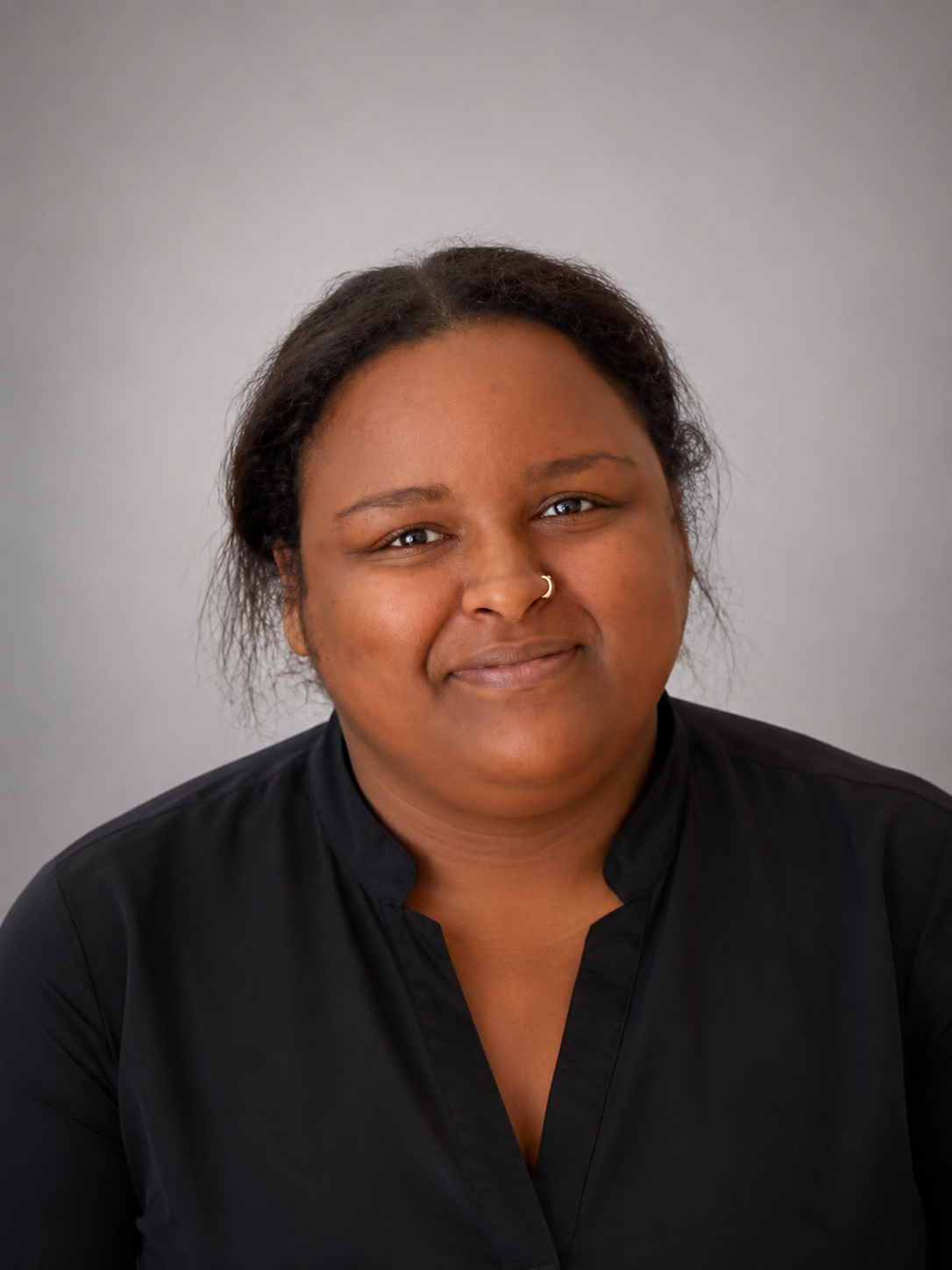 A woman with medium skin tone, dark curly hair, and a nose ring, wearing a black shirt, smiling at the camera against a plain gray background.