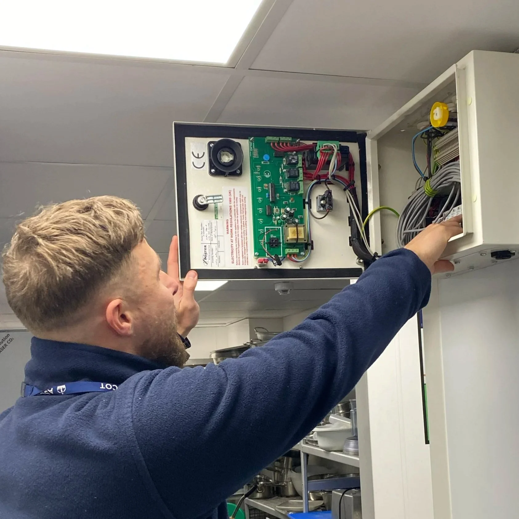 A person working on an electrical control panel in a commercial kitchen or industrial setting, with shelves of kitchen equipment and a ceiling light visible.