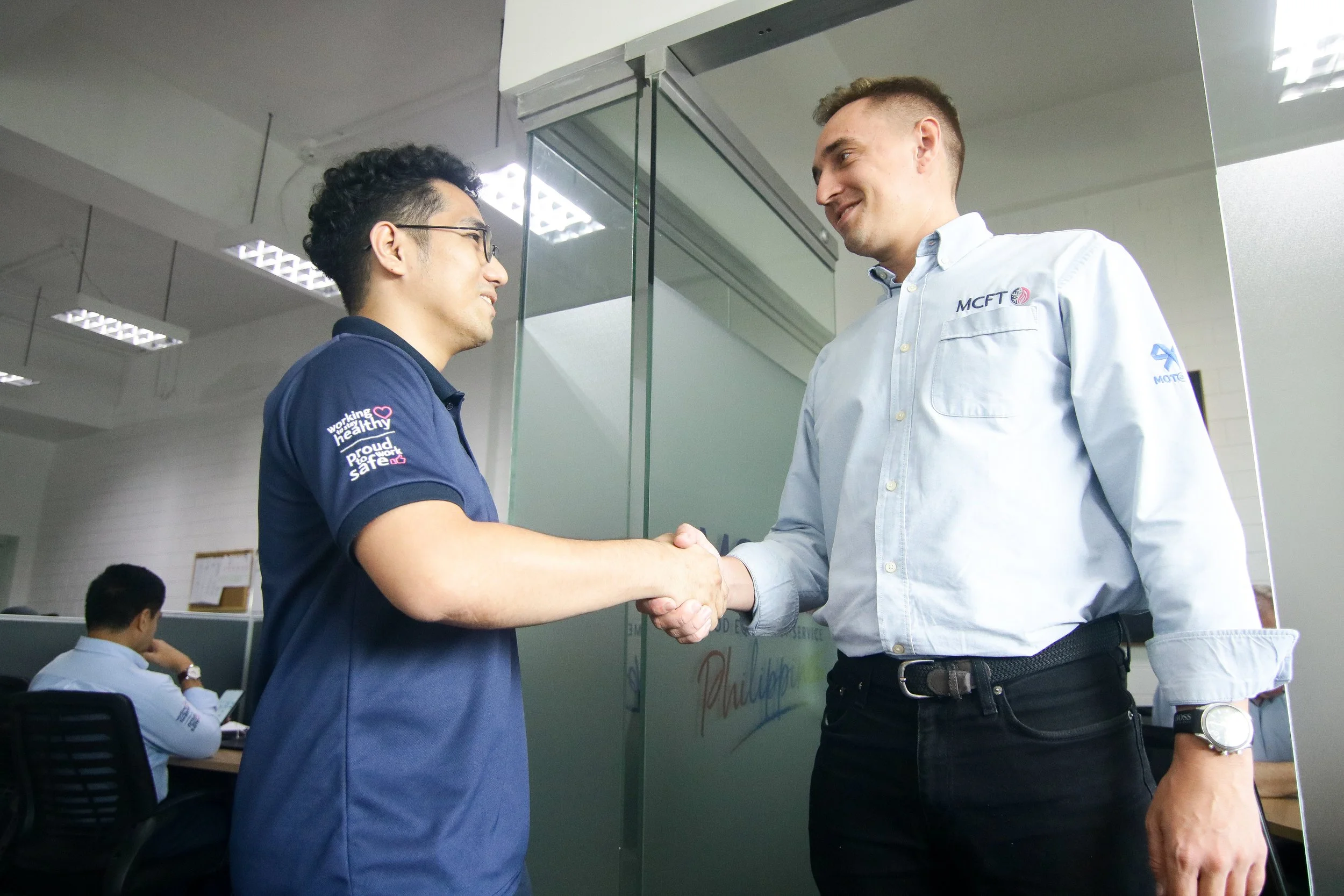 Two men shaking hands in an office, smiling at each other, one wearing a blue shirt and glasses, the other in a white shirt with company logos, in front of a glass wall.