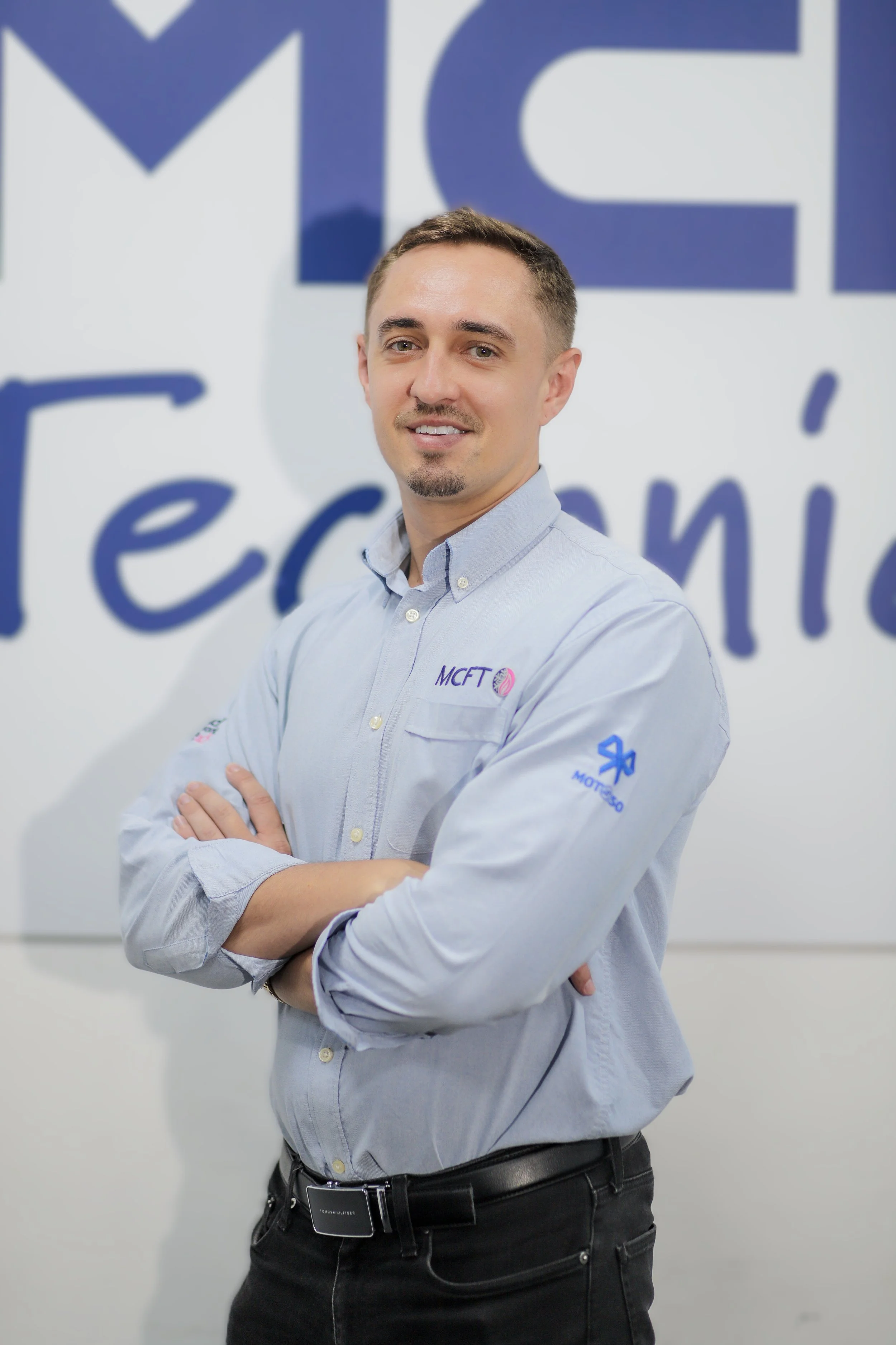 A man with short brown hair, a trimmed beard, wearing a light blue button-up shirt with company logos, standing with arms crossed in front of a large sign with blue and white text, smiling at the camera.