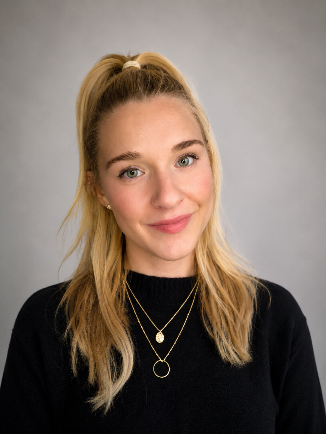 A young woman with blonde hair in a high ponytail, wearing a black top and layered gold necklaces, smiling at the camera against a gray background.