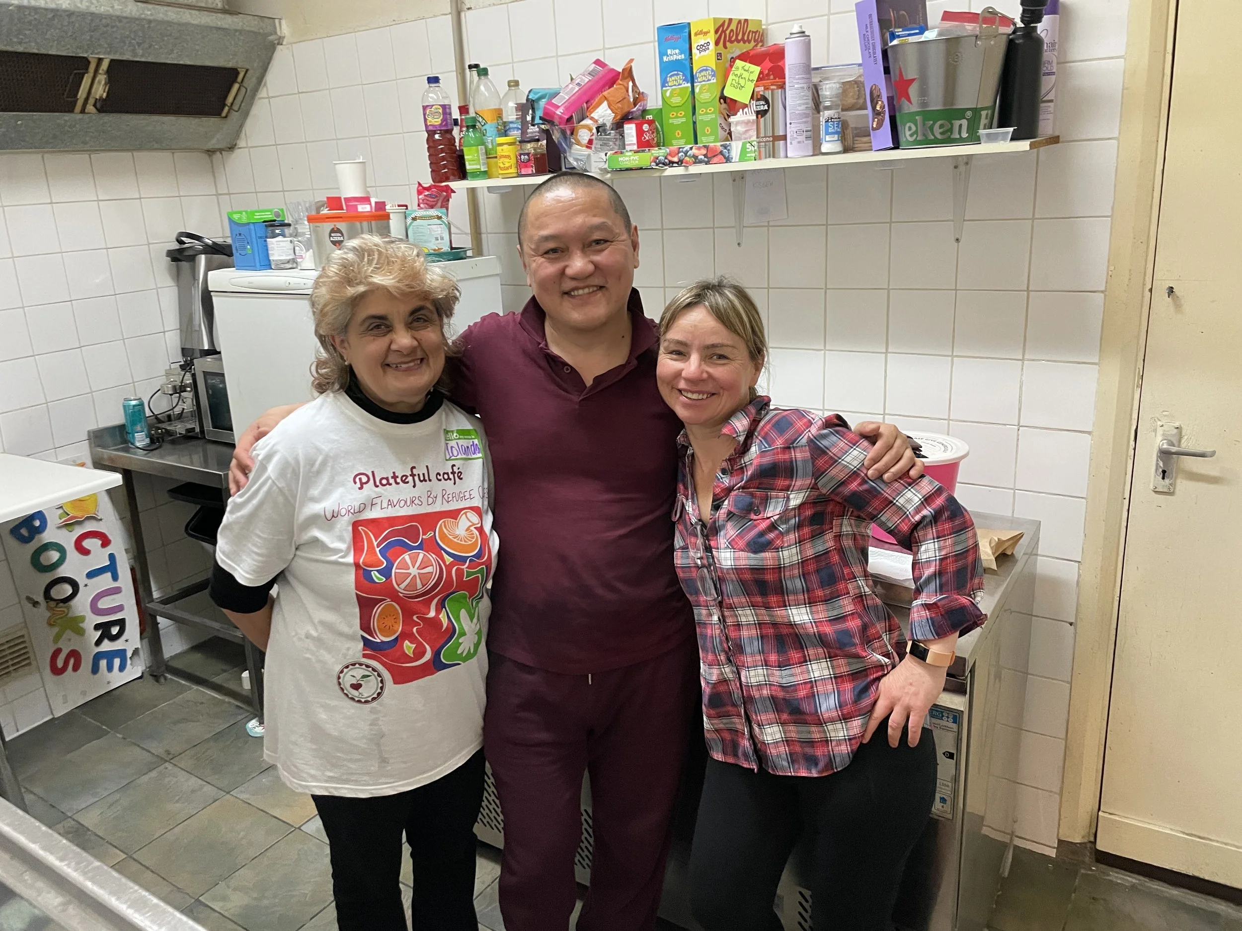 Three people smiling and hugging in a kitchen with shelves of food and a white tiled wall in the background.