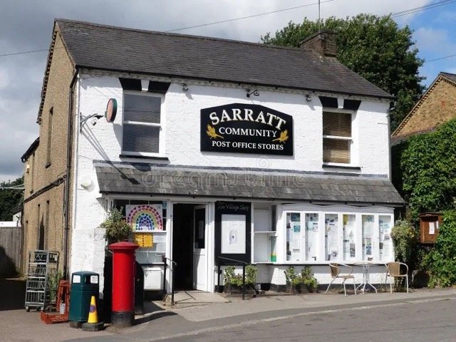 Small white building with a sign reading "Sarratt Community Post Office Stores." Outside, there are a red post box, a black mailbox, recycling bins, and a few chairs. The building has two upper windows and a slate roof.