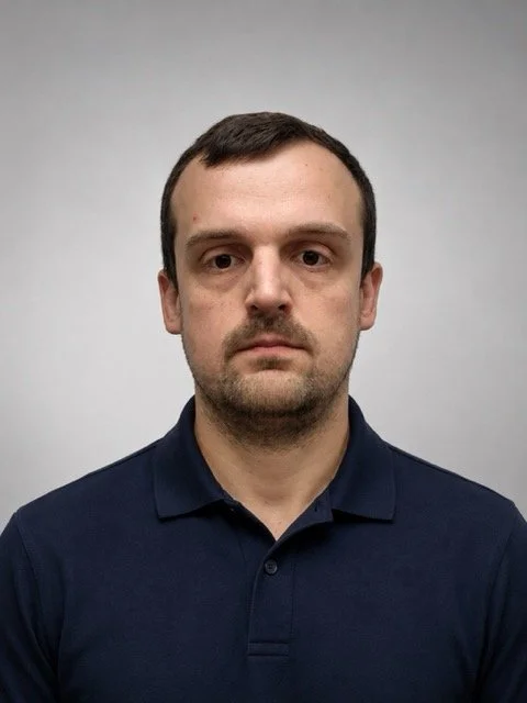 A man with short dark hair, light skin, wearing a navy blue polo shirt against a plain light gray background.