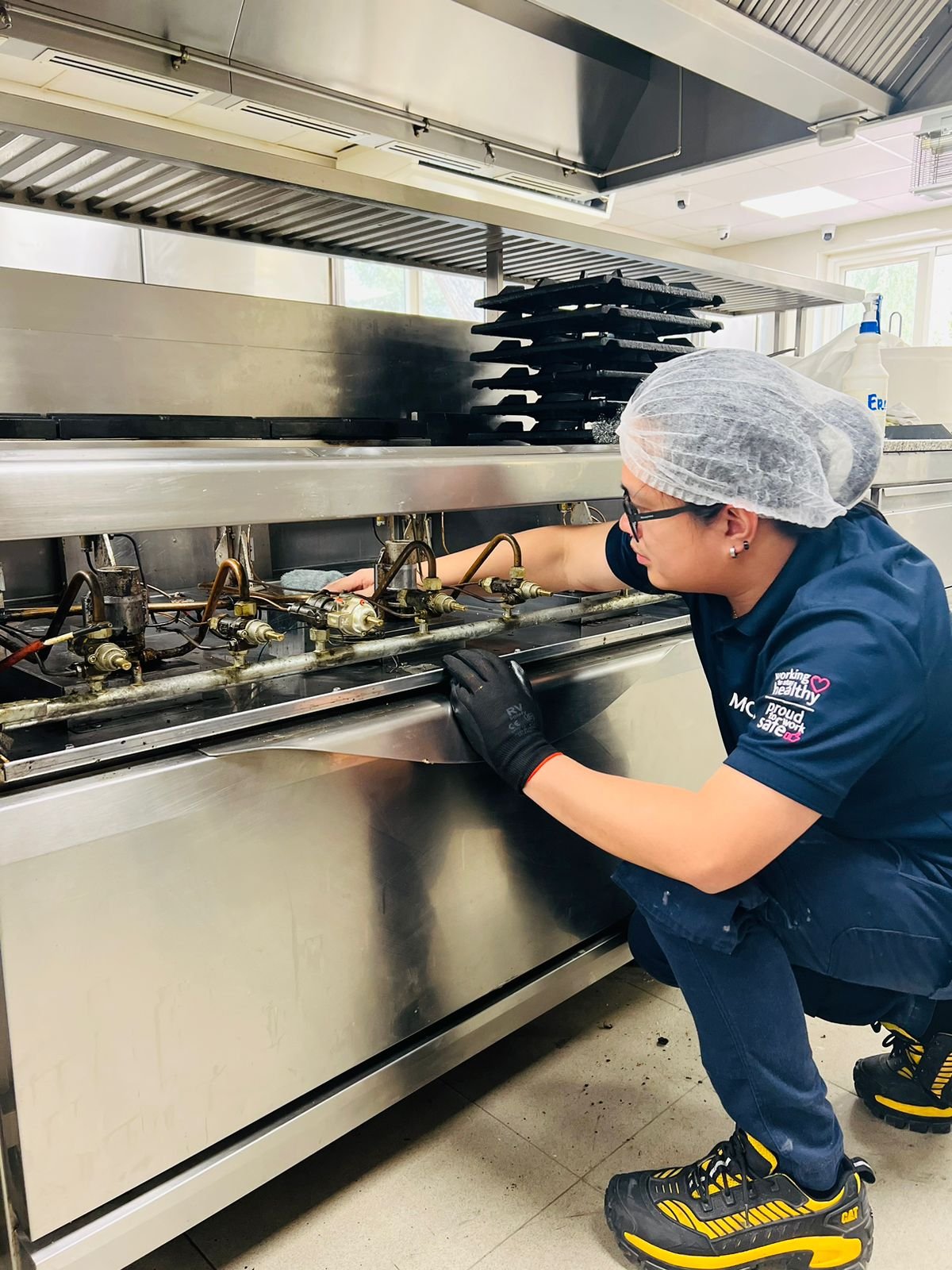 A person wearing a hairnet, glasses, and gloves inspecting or repairing a commercial kitchen stove.