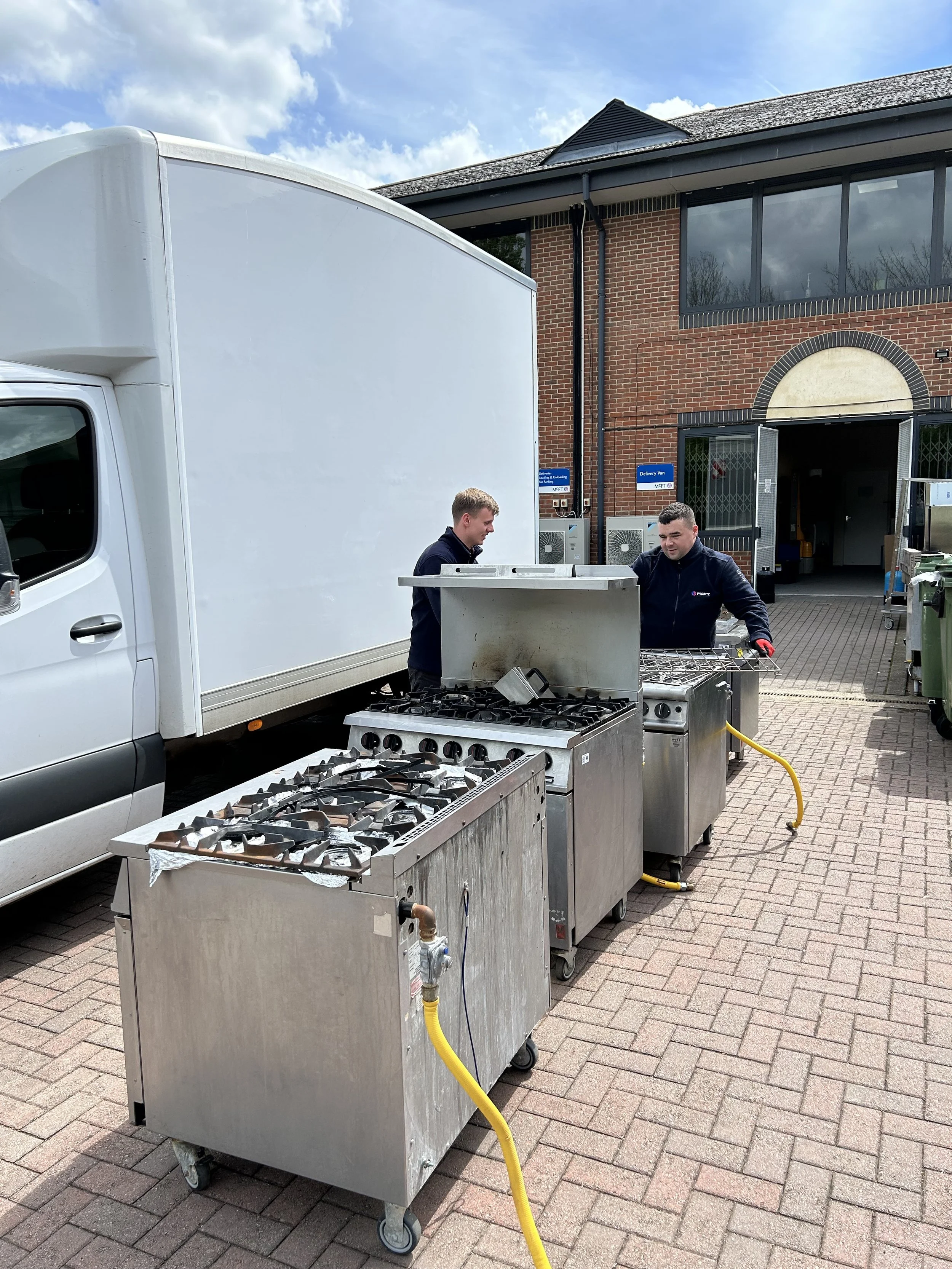 Two workers set up outdoor propane fryers near a delivery van outside a brick building with large windows.