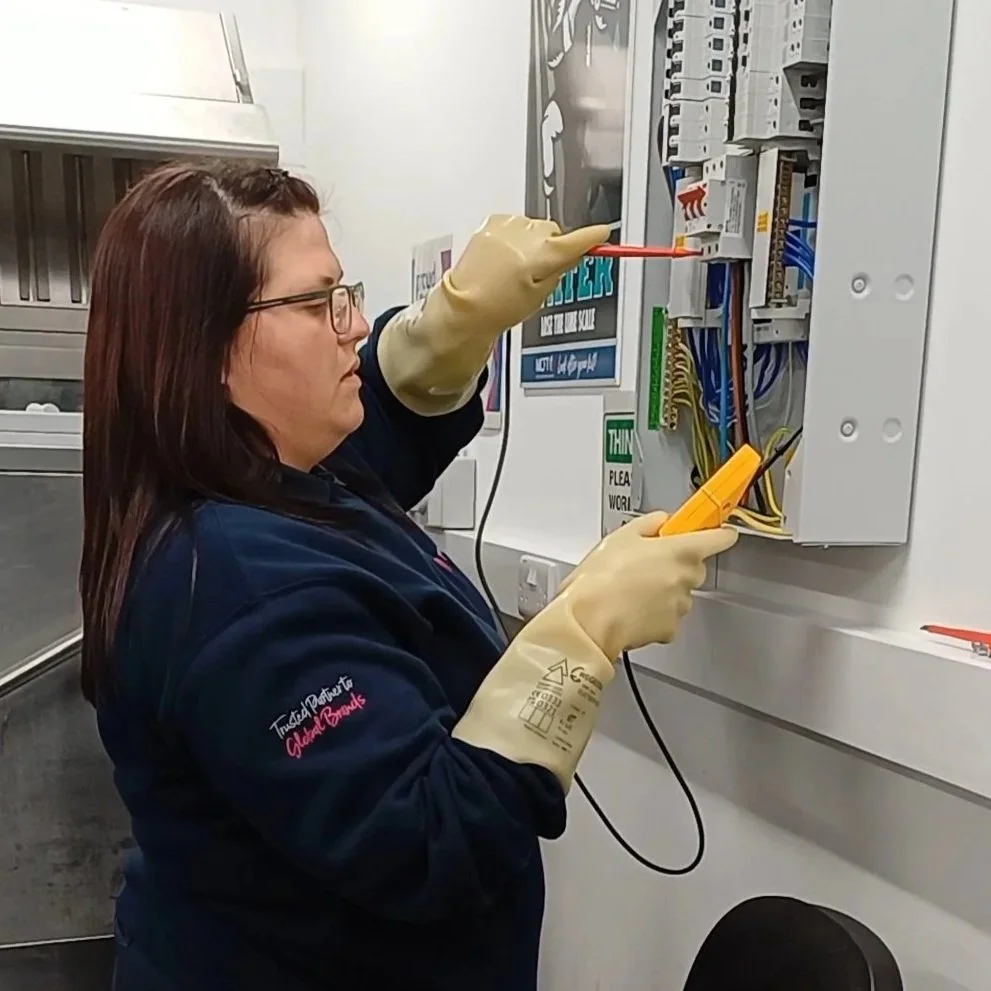 A woman working on an electrical panel, using tools and wearing rubber gloves.
