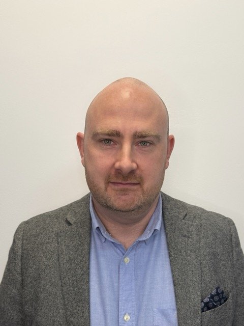 A bald man with a beard wearing a gray blazer and blue collared shirt, standing against a plain white wall.